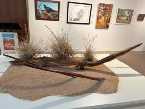 A mixed media artwork showing a carved goanna and boomerangs on a spinifex landscape.